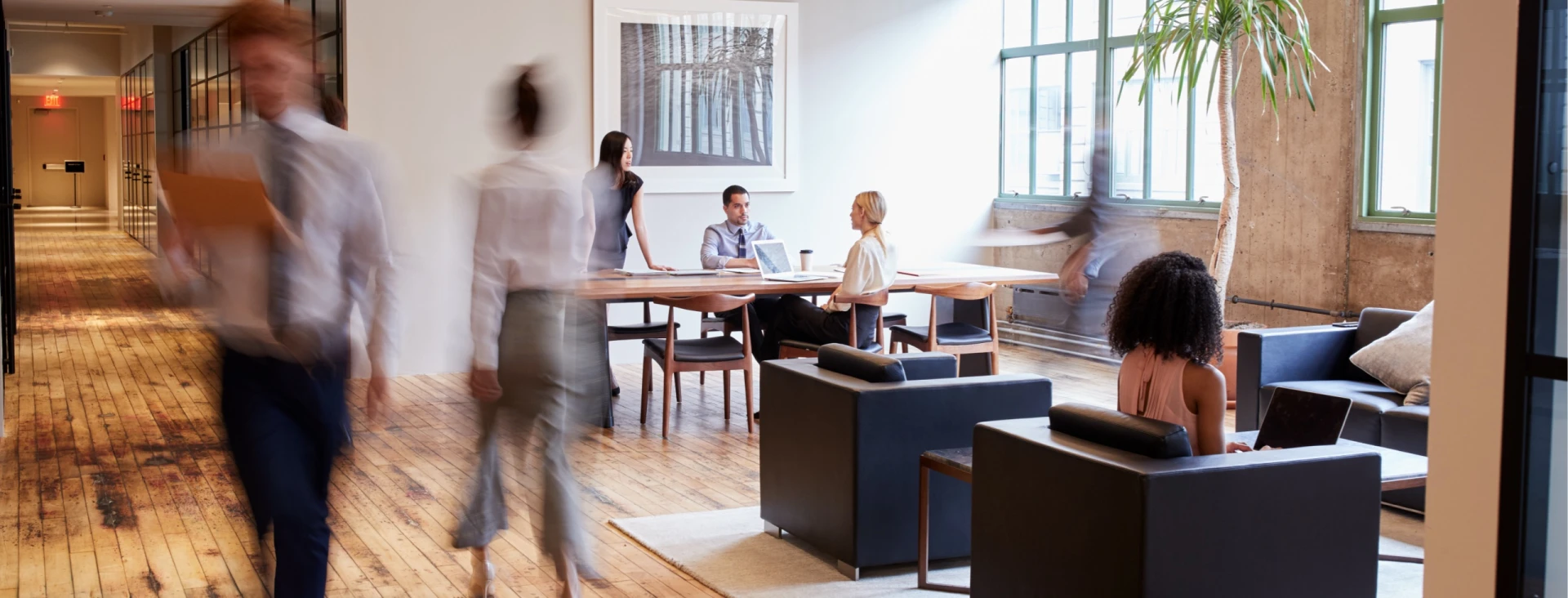 A group of people sitting at a table in an office, some engrossed in conversation while others are focused on their laptops.