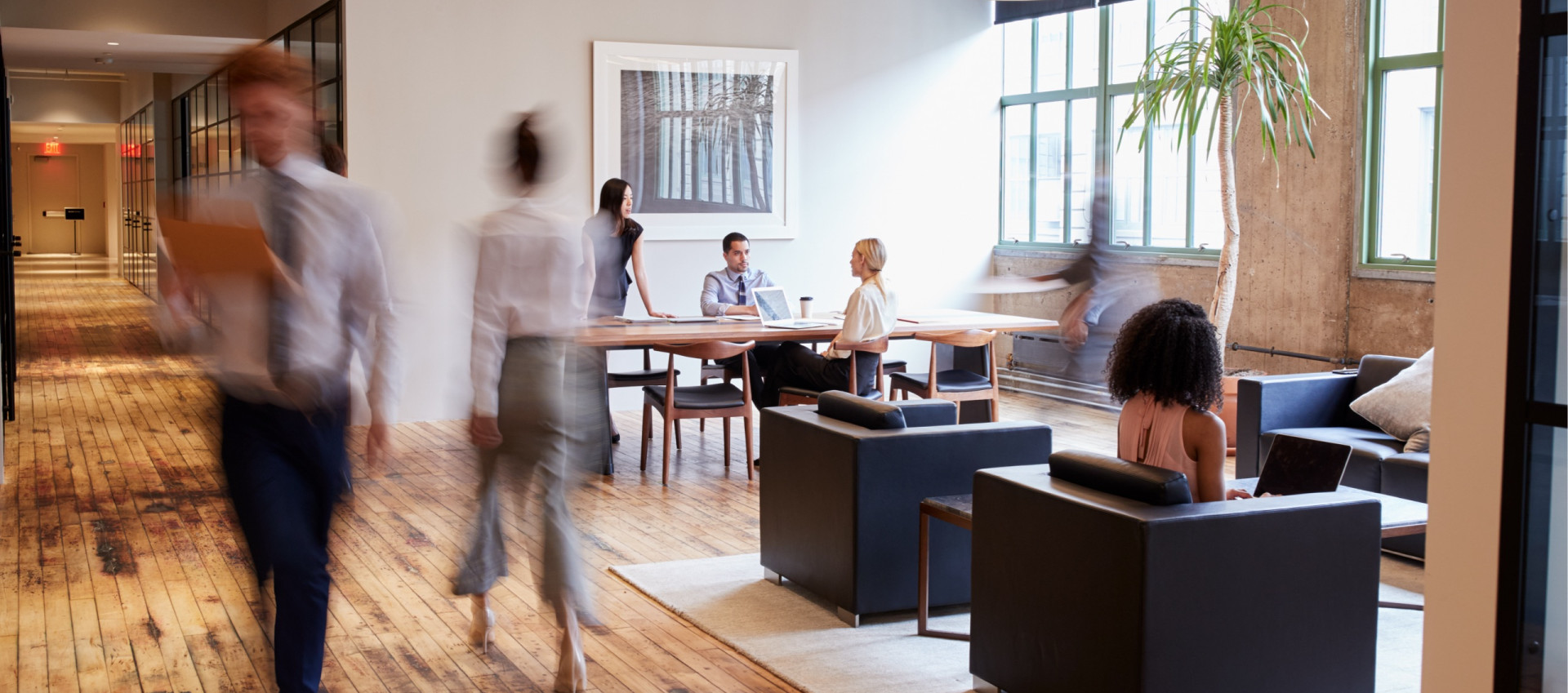 A group of people sitting at a table in an office, some engrossed in conversation while others are focused on their laptops.