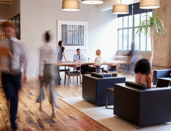 A group of people sitting at a table in an office, some engrossed in conversation while others are focused on their laptops.