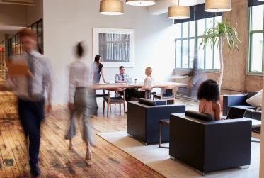 A group of people sitting at a table in an office, some engrossed in conversation while others are focused on their laptops.