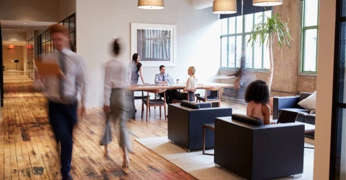 A group of people sitting at a table in an office, some engrossed in conversation while others are focused on their laptops.
