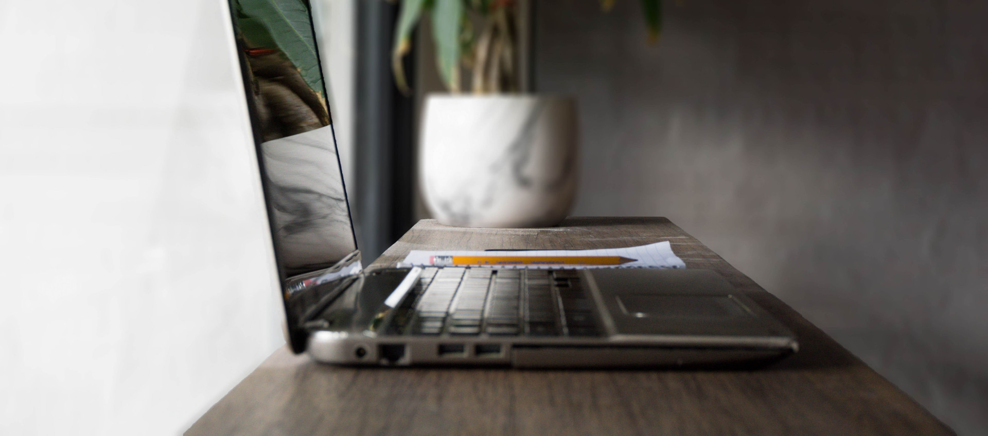 side view of laptop, notepad and pencil on a skinny wooden table with blurred out plant in background