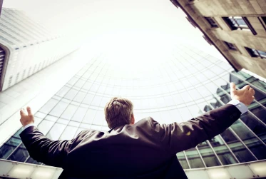 low angle view of man in suit with arms out wide towards the sky in front of high-rise buildings
