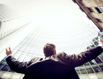 low angle view of man in suit with arms out wide towards the sky in front of high-rise buildings