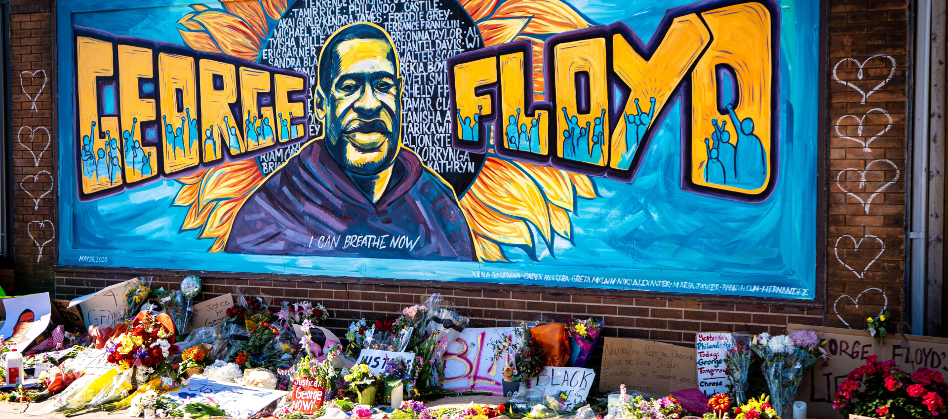 george floyd memorial on a brick wall with several flowers and messages on the sidewalk.