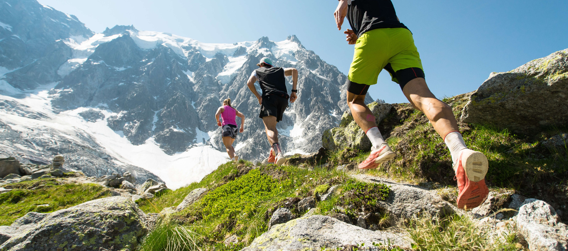 low angle view of three people running across rocks towards a snow-covered mountain