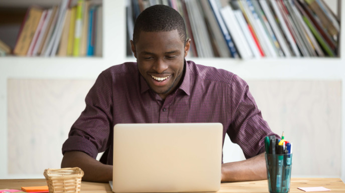 man smiling and sitting at a desk with laptop, pens and post-it notes on the table