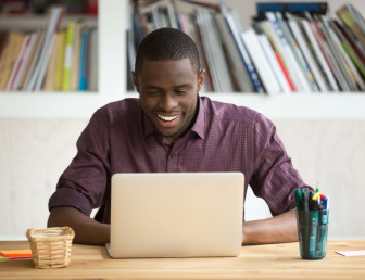 man smiling and sitting at a desk with laptop, pens and post-it notes on the table