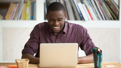 man smiling and sitting at a desk with laptop, pens and post-it notes on the table