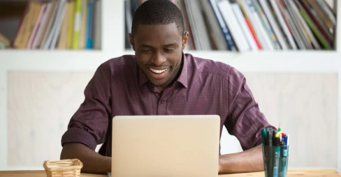 man smiling and sitting at a desk with laptop, pens and post-it notes on the table