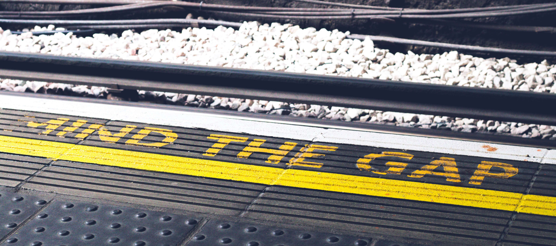 the word "mind the gap" in yellow writing on the floor with electric train wiring in the background
