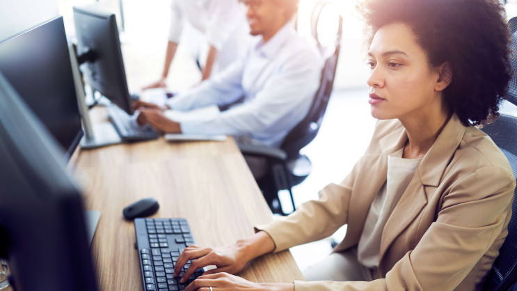 side view of woman in smart clothing working at a desk with two people blurred in the background