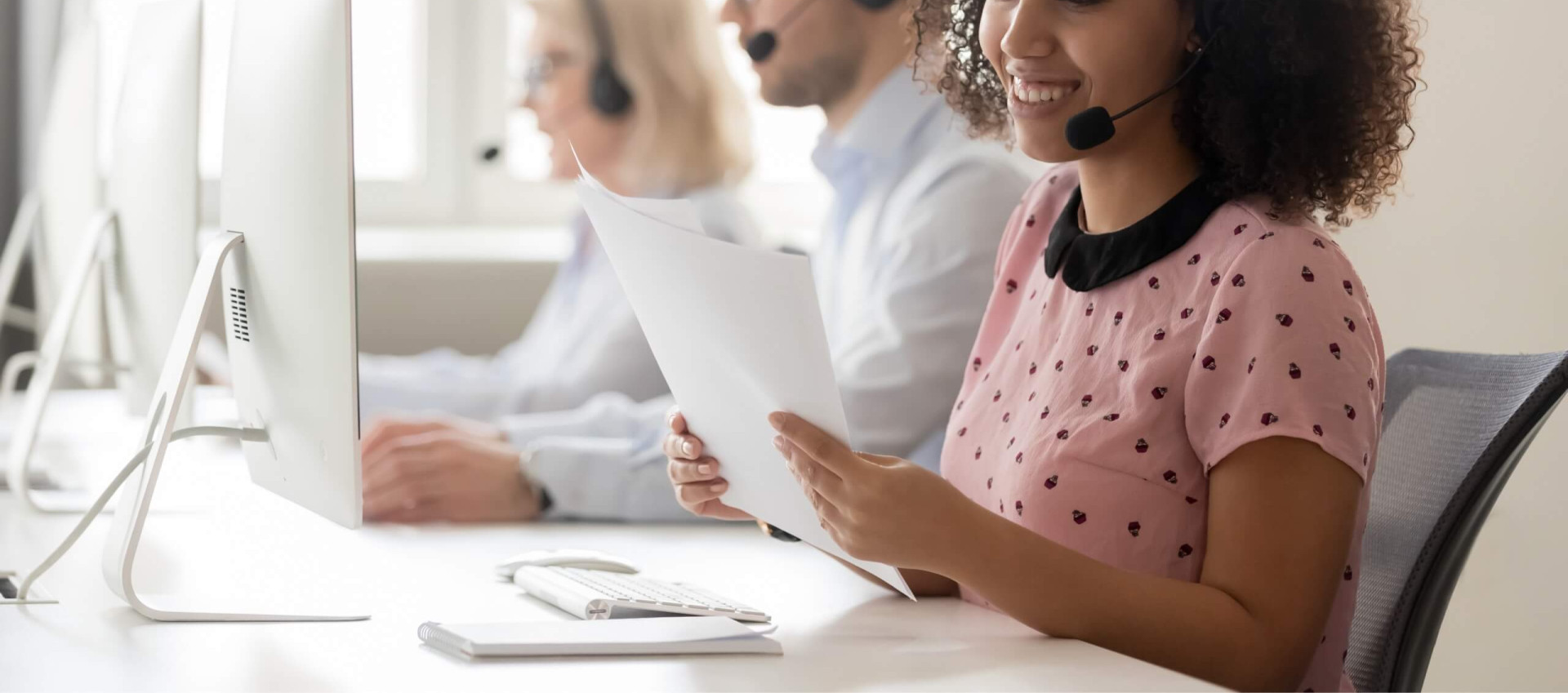 woman at a desk in a call center with headset on holding documents with two people in background