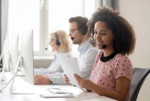 woman at a desk in a call center with headset on holding documents with two people in background