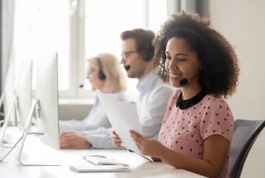 woman at a desk in a call center with headset on holding documents with two people in background