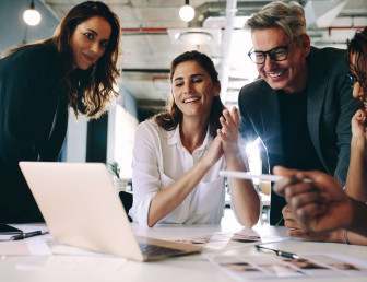 Four colleagues in an office, all smiles, gathered around a laptop screen.