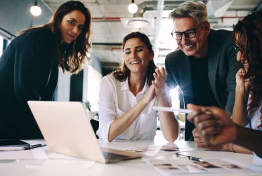 Four colleagues in an office, all smiles, gathered around a laptop screen.