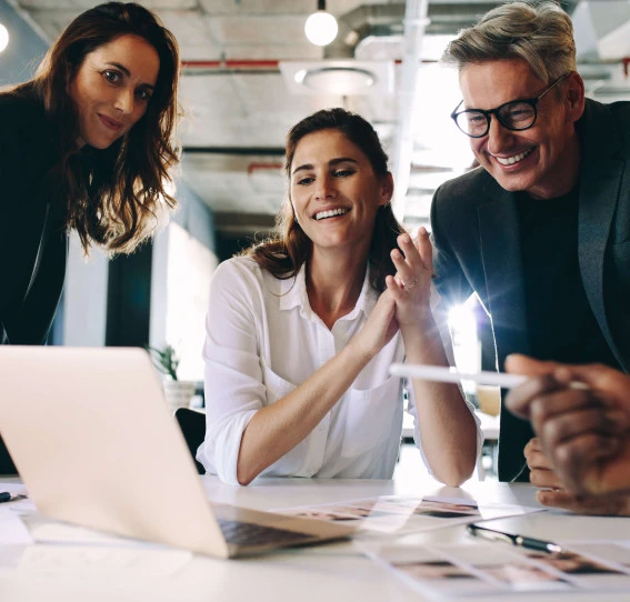 Four colleagues in an office, all smiles, gathered around a laptop screen.