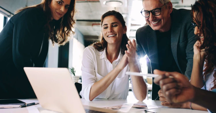 Four colleagues in an office, all smiles, gathered around a laptop screen.