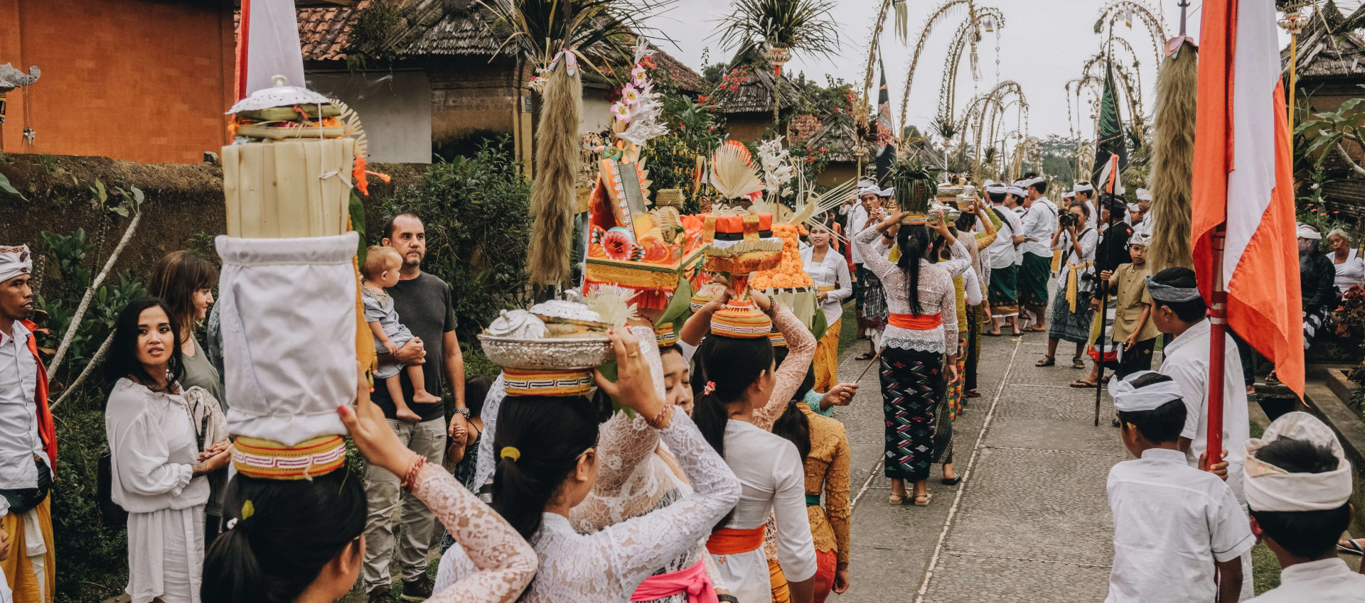 several people walking one by one holding baskets of items on their heads with bystanders watching