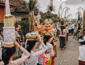 several people walking one by one holding baskets of items on their heads with bystanders watching