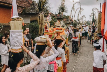 several people walking one by one holding baskets of items on their heads with bystanders watching