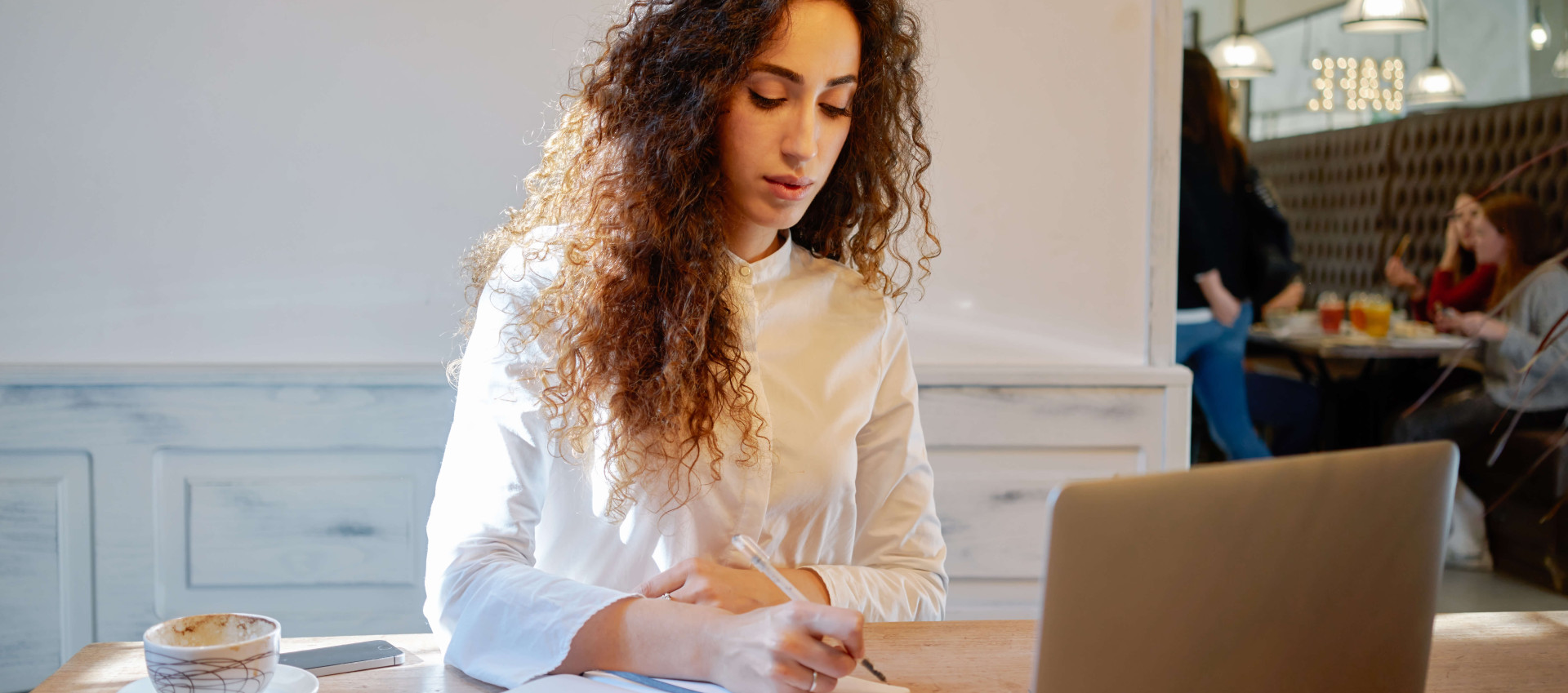 eye level view of woman in café writing in notebook with coffee and laptop on table