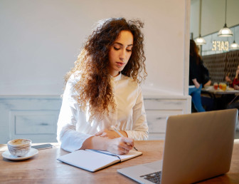 eye level view of woman in café writing in notebook with coffee and laptop on table