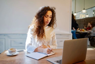 eye level view of woman in café writing in notebook with coffee and laptop on table