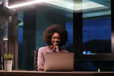 woman working in the evening smiling and sitting at a desk in an office with a laptop