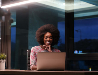 woman working in the evening smiling and sitting at a desk in an office with a laptop