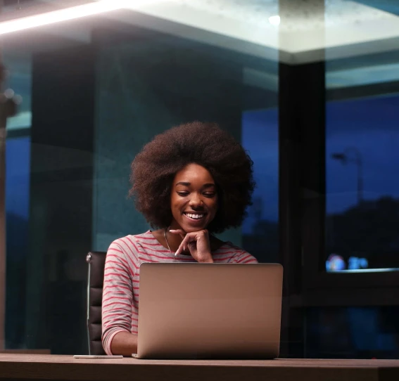 woman working in the evening smiling and sitting at a desk in an office with a laptop