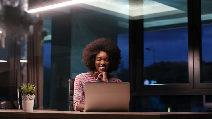 woman working in the evening smiling and sitting at a desk in an office with a laptop