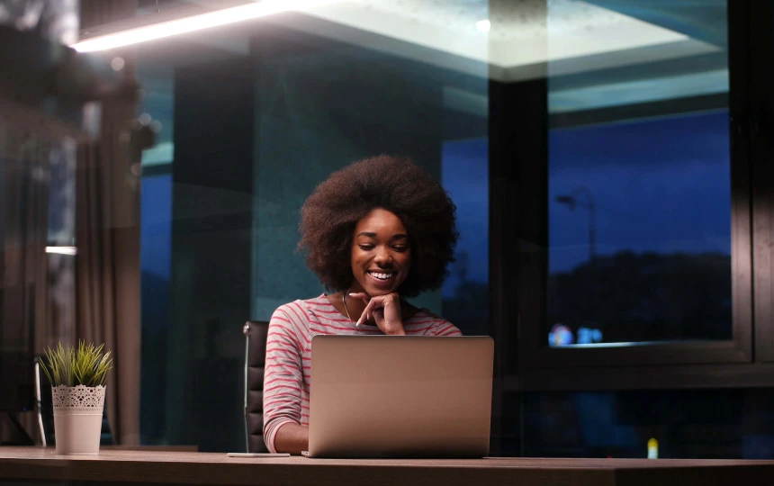 woman working in the evening smiling and sitting at a desk in an office with a laptop