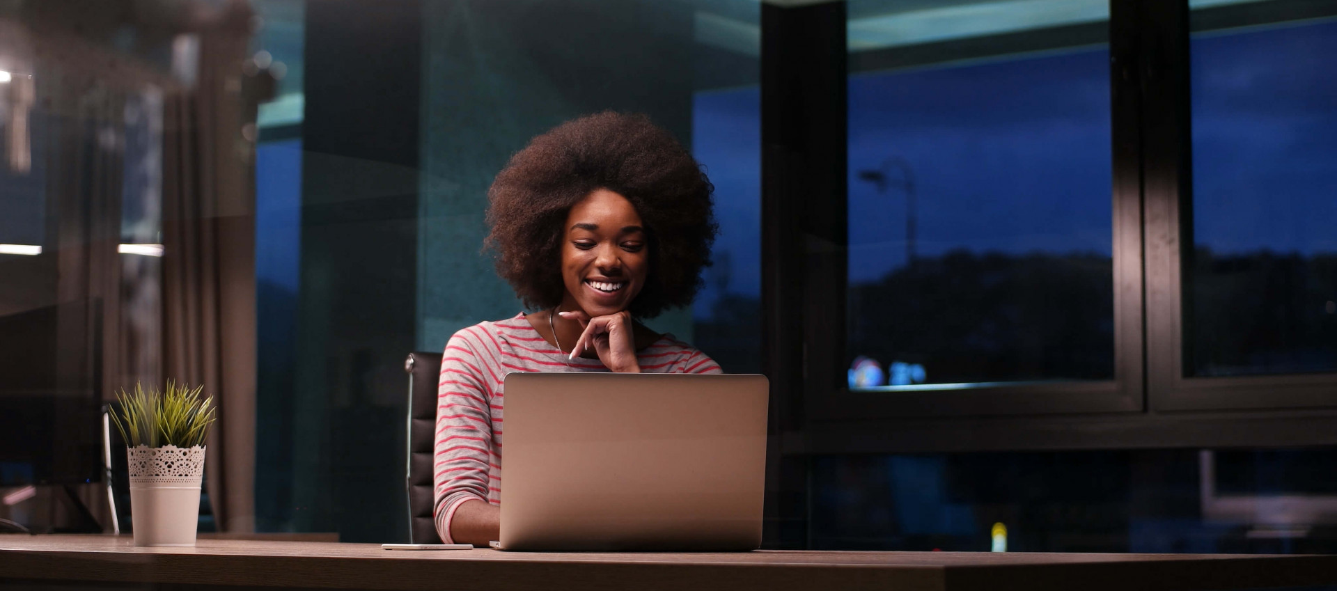 woman working in the evening smiling and sitting at a desk in an office with a laptop