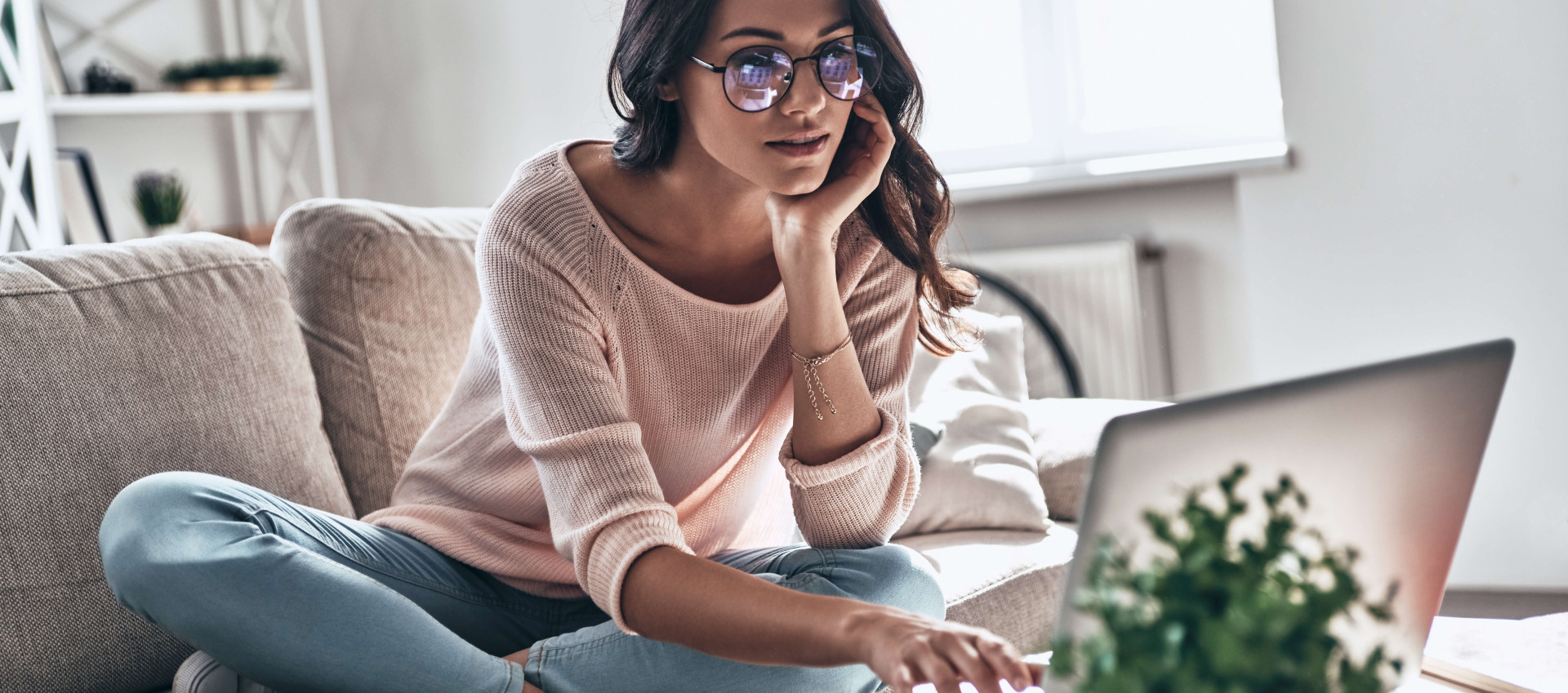 woman with glasses wearing casual clothes sitting with legs crossed on sofa looking at a laptop