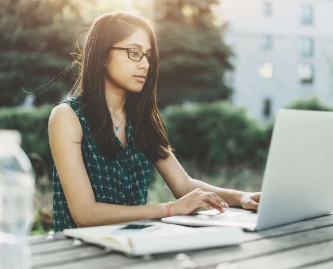 eye level view of woman sitting outside on bench table with laptop, notepad and bottle of water
