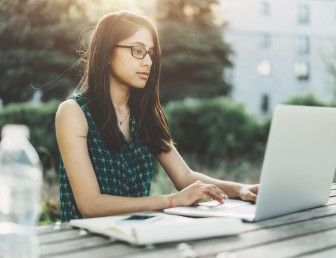eye level view of woman sitting outside on bench table with laptop, notepad and bottle of water