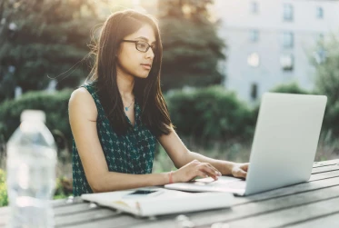 eye level view of woman sitting outside on bench table with laptop, notepad and bottle of water