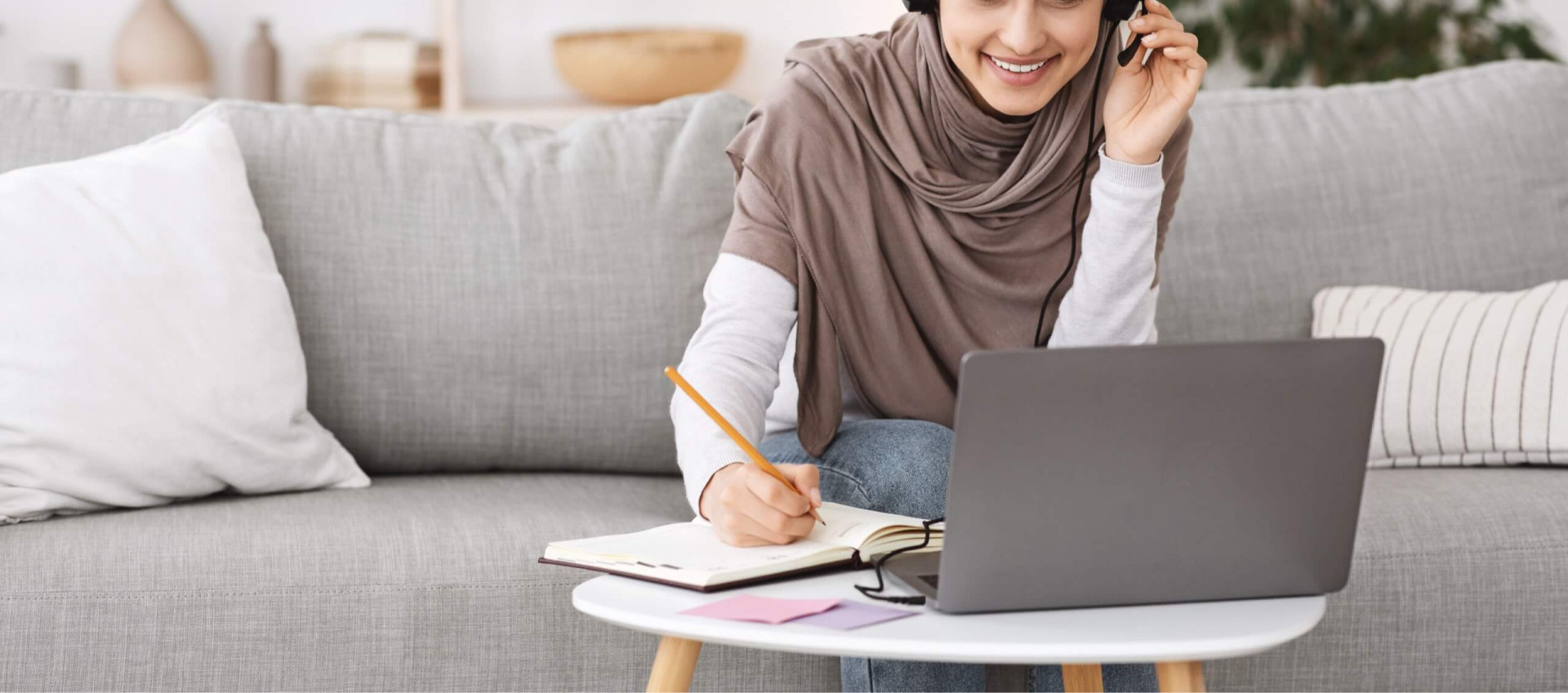 woman sitting on sofa wearing hijab and casual clothes working from home with headset and laptop