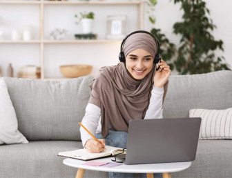 woman sitting on sofa wearing hijab and casual clothes working from home with headset and laptop