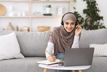woman sitting on sofa wearing hijab and casual clothes working from home with headset and laptop