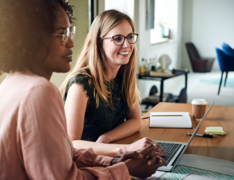 eye level view of two women at a table with laptop in front speaking to others out of view