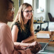 eye level view of two women at a table with laptop in front speaking to others out of view