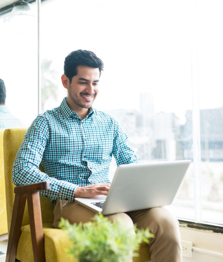 man sitting in an armchair next to window with laptop on his knees 