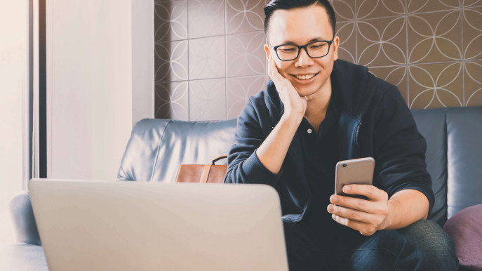 front view of man in causal clothes sitting on sofa smiling and holding mobile phone with laptop in view