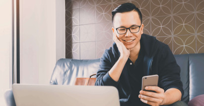 front view of man in causal clothes sitting on sofa smiling and holding mobile phone with laptop in view
