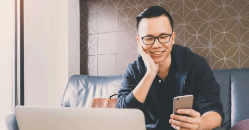 front view of man in causal clothes sitting on sofa smiling and holding mobile phone with laptop in view