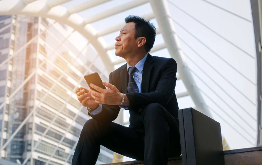 Asian man in a suit sitting on steps with a brief case and phone in hand looking up to the sky