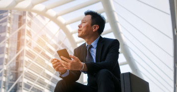 Asian man in a suit sitting on steps with a brief case and phone in hand looking up to the sky